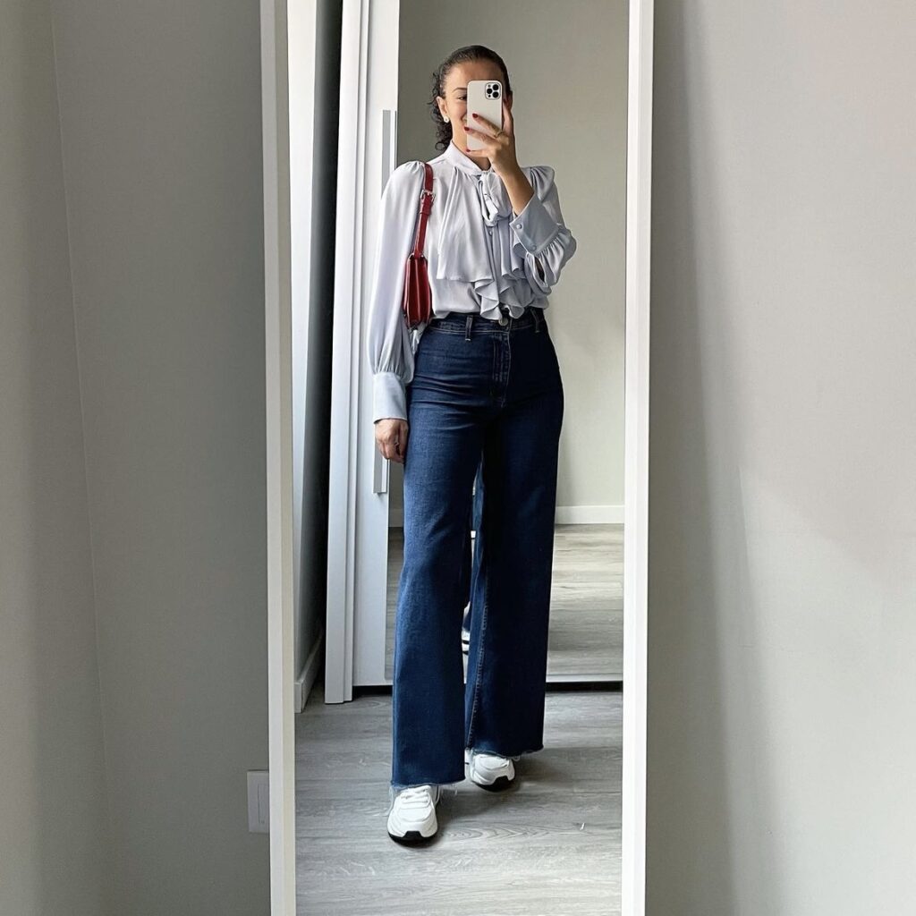 photo of woman with ruffled top, dark denim jeans, and trainers