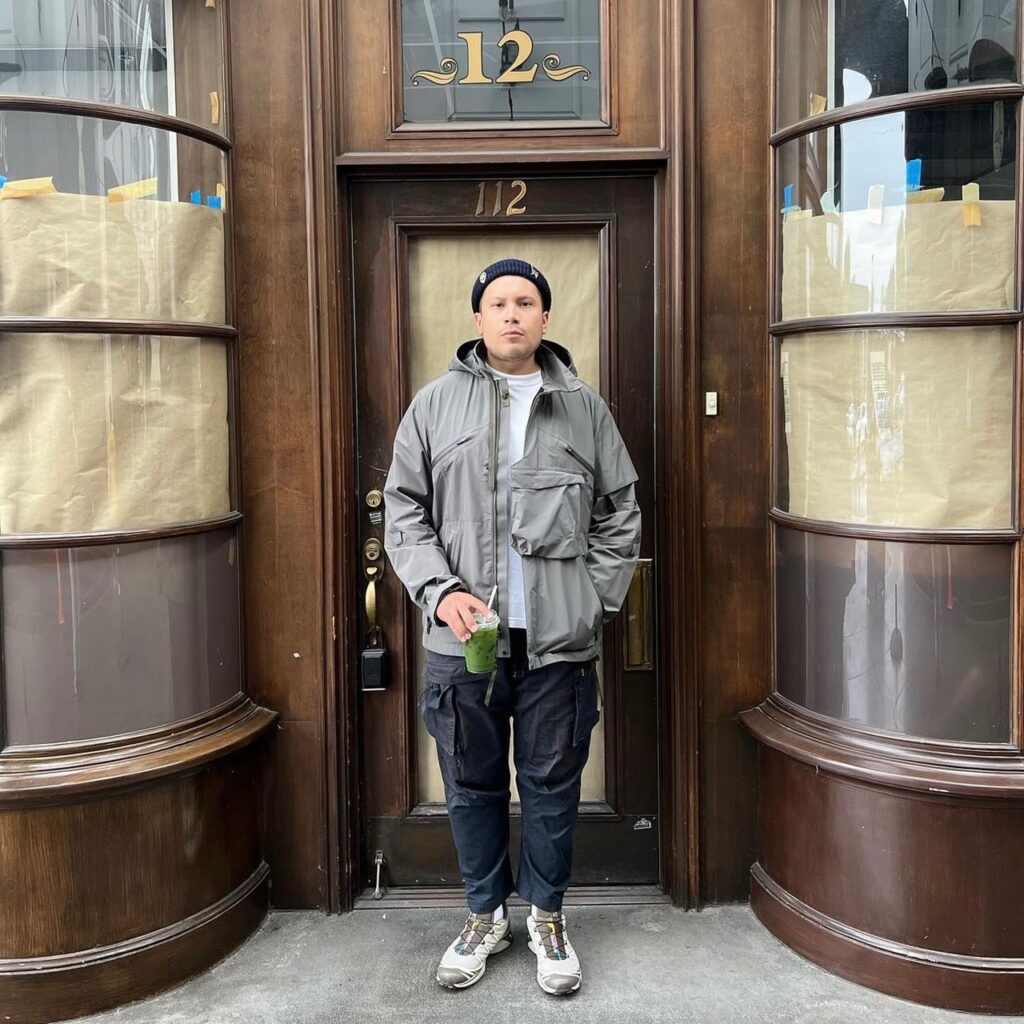 man wearing a rain jacket and cargo pants with hiking shoes, standing in front of a cafe